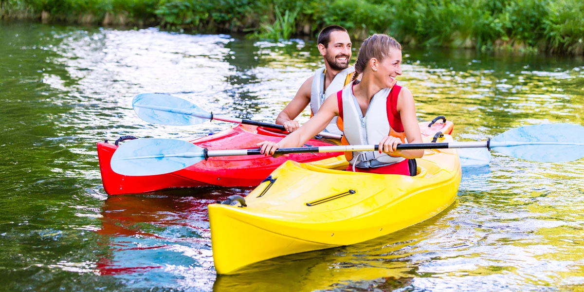Paar beim Kajakfahren auf einem Fluss: Frau im gelben Kajak und Mann im roten Kajak mit Paddeln und Schwimmwesten.