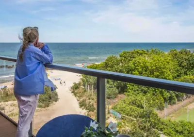 Frau auf der Terrasse mit Blick auf Ostsee, Strandzugang und grüne Dünenlandschaft bei sonnigem Wetter.