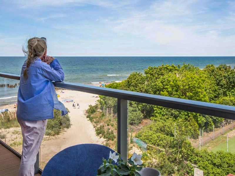 Frau auf der Terrasse mit Blick auf Ostsee, Strandzugang und grüne Dünenlandschaft bei sonnigem Wetter.