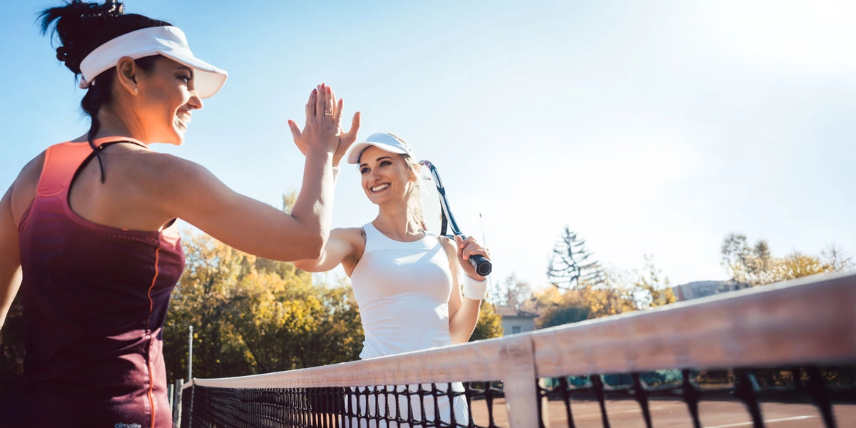 Ein sonniger Tag auf dem Tennisplatz, zwei Tennisspielerinnen geben sich am Netz ein High-Five, eine hält den Schläger.
