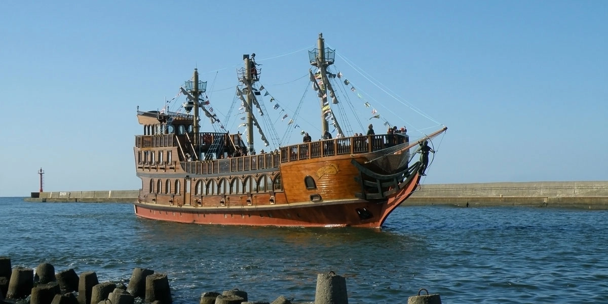 Touristisches Holzschiff mit Flaggen vor Dziwnóws Hafeneinfahrt und Ostsee-Panorama im Hintergrund