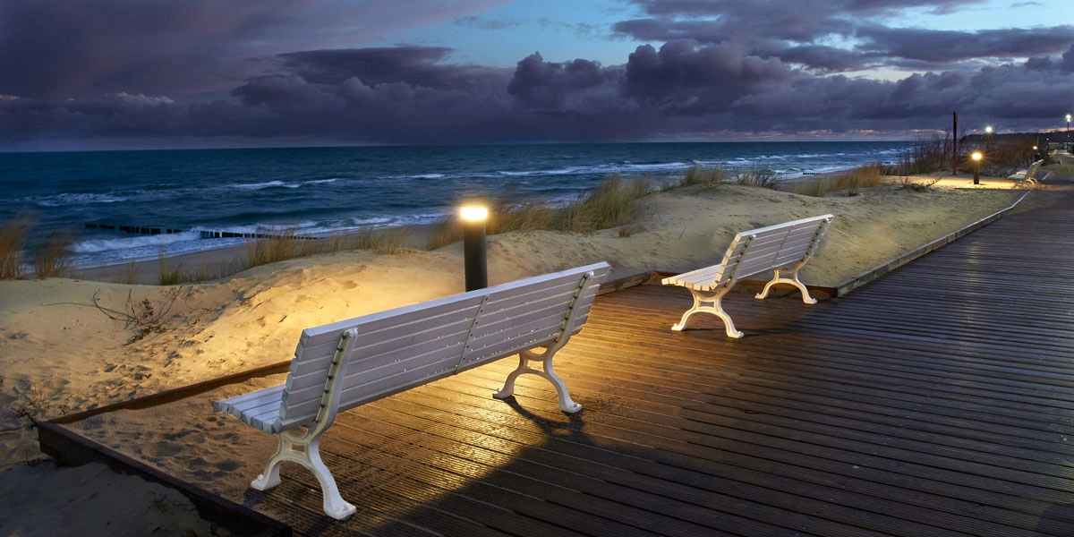 Beleuchtete Strandpromenade in Dziwnów bei Abenddämmerung mit Bänken, Dünen und Blick auf die Ostsee.