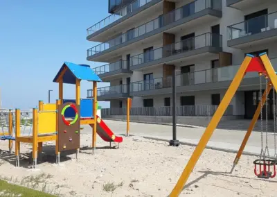 Spielplatz neben der Ferienanlage MorSky Gardenia in Dziwnów mit Klettergerüst, Rutsche und Schaukel vor dem Gebäude.