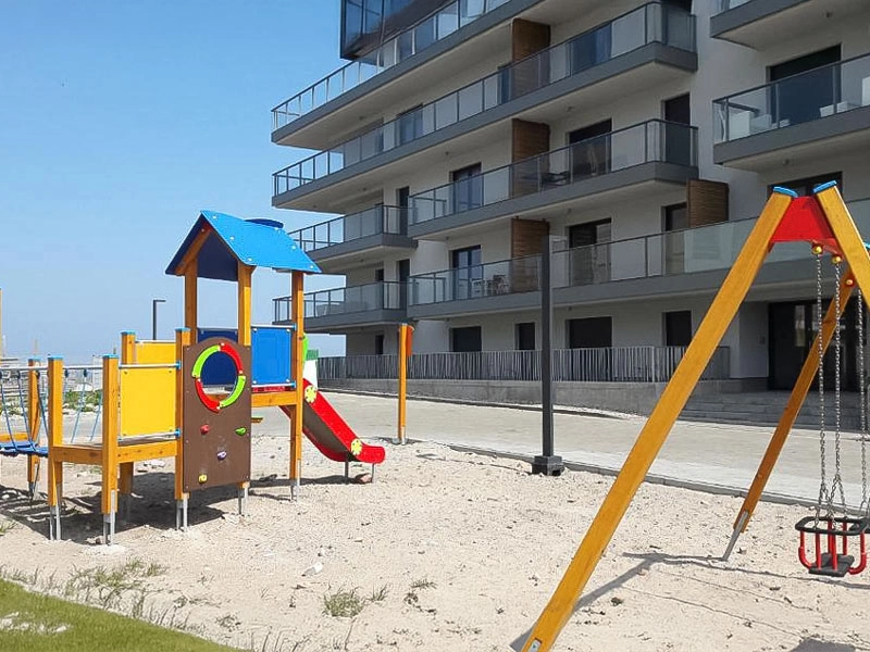 Spielplatz neben der Ferienanlage MorSky Gardenia in Dziwnów mit Klettergerüst, Rutsche und Schaukel vor dem Gebäude.