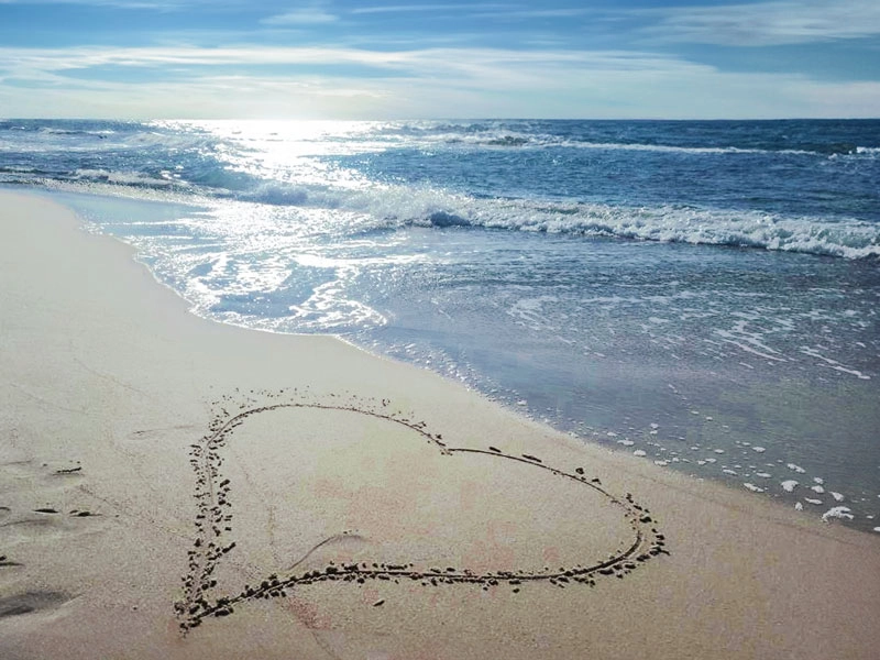 Herz im Sand am Ostseestrand, sanfte Wellen im Gegenlicht bei ruhiger Stimmung am Meer.